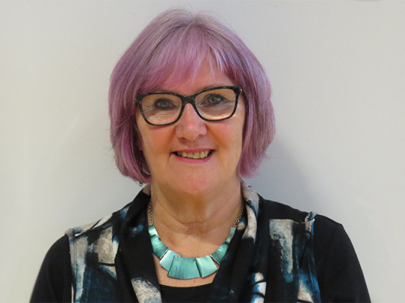 A woman with short pink hair is standing against a grey background and smiling.