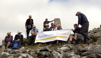 Eleven people standing at the top of a mountain (Brown Willy) next to a marker stone, holding up a sign that says Tiverton u3a