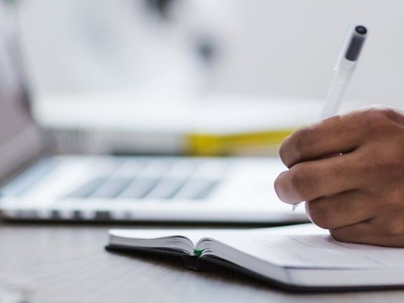 person holding pen writing notes next to laptop